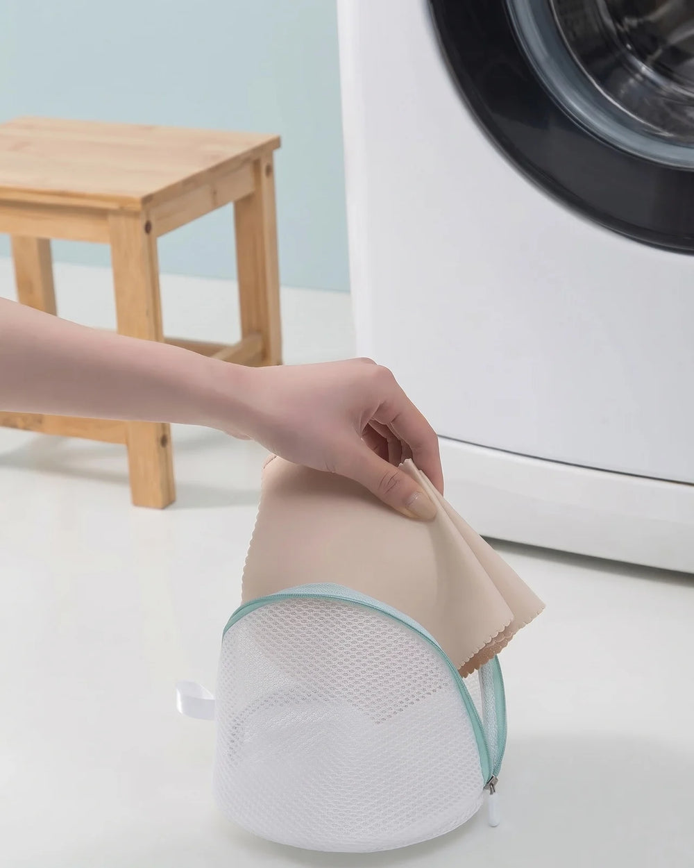 Hand lowering a beige garment into the cylindrical mesh wash bag beside a washing machine